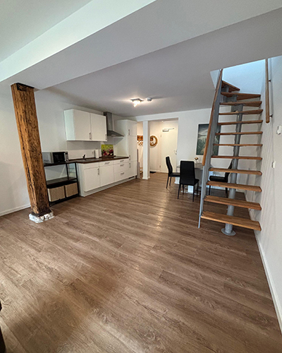 Offene Küche mit weißem Schrank; Holztreppe, Holzboden, schwarzer Esstisch; heller Raum.