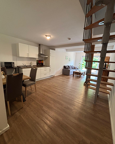 Helles Wohnzimmer mit Holzboden, moderner Küche, grauen Stühlen, Sofa und Wendeltreppe.