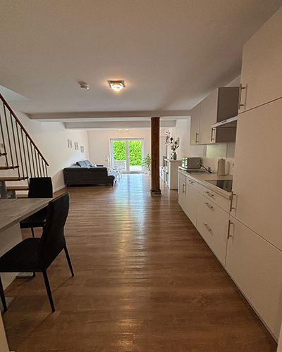Helles Wohnzimmer mit Holzfußboden, Sofa, Esstisch, offener Küche und Blick auf grüne Hecke.